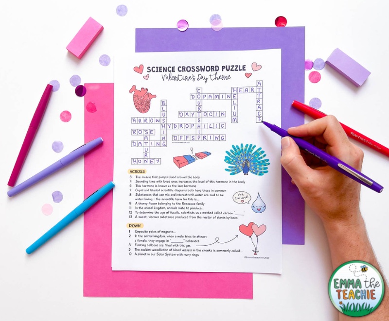 An overhead view of a science crossword puzzle with a Valentine's Day theme against pink and purple card. A hand is holding a flair pen and writing answers on the crossword. There are other pens, colored confetti and mini post-its around the puzzle.