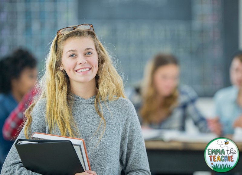 An image of a happy female student that is holding a binder and books, with other students in the background.