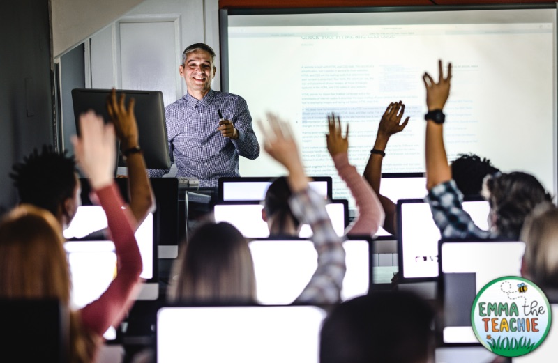 An image of a teacher at the front of the room in front of a class of students with all of their hands up ready to participate.