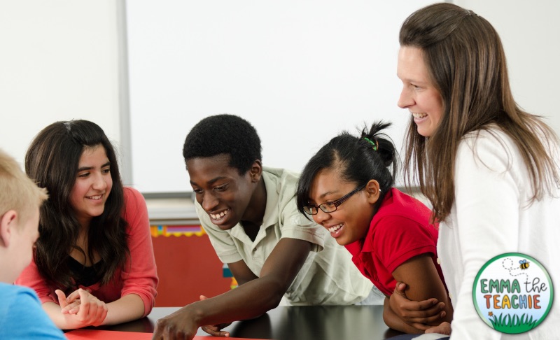 An image of a teacher overseeing a group of students working together. They are all smiling and engaged in the activity on the desk.