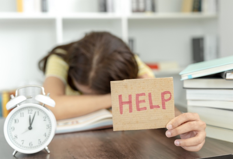 An image of a student with her head on the desk surrounded by a clock and books. She is holding a sign that says, “HELP”!