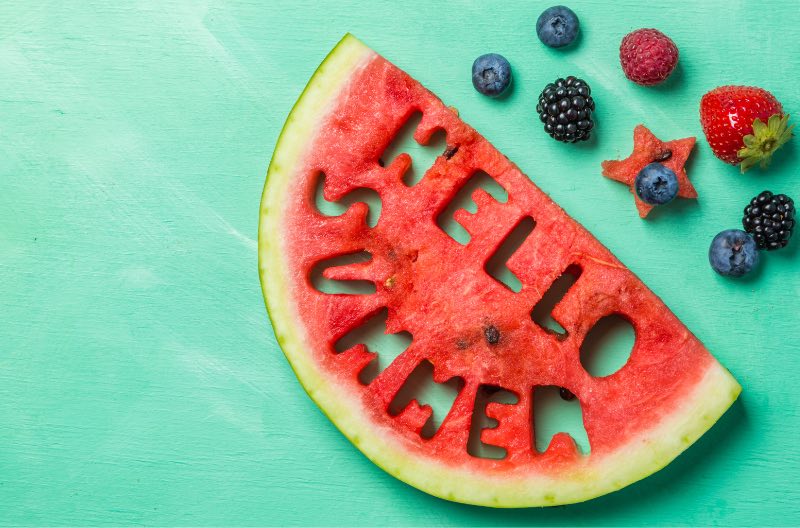 An image with “HELLO SUMMER” cut out of a half watermelon. There are berries laid around the watermelon.