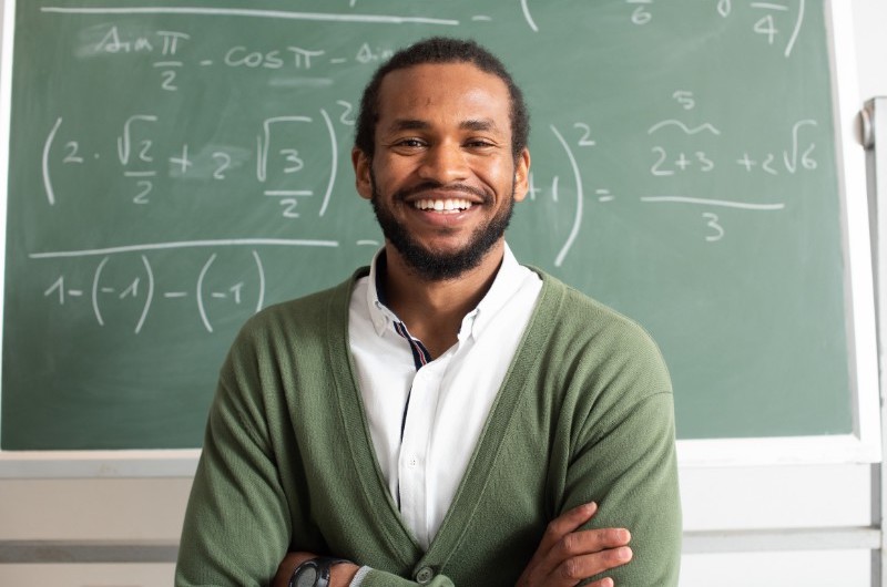 An image of a smiling male teacher standing in front of a board.