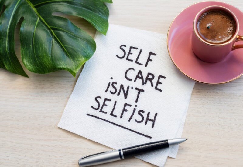 An image of a table with a plant in the upper left, a cup of coffee in the upper right, and a piece of paper with a pen in the center. The piece of paper reads, “Self care isn’t selfish.”