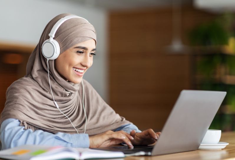 A young woman is smiling while using her laptop. She is typing and wearing headphones. This is to show that professional development for science teachers can be through online courses like National Geographic.