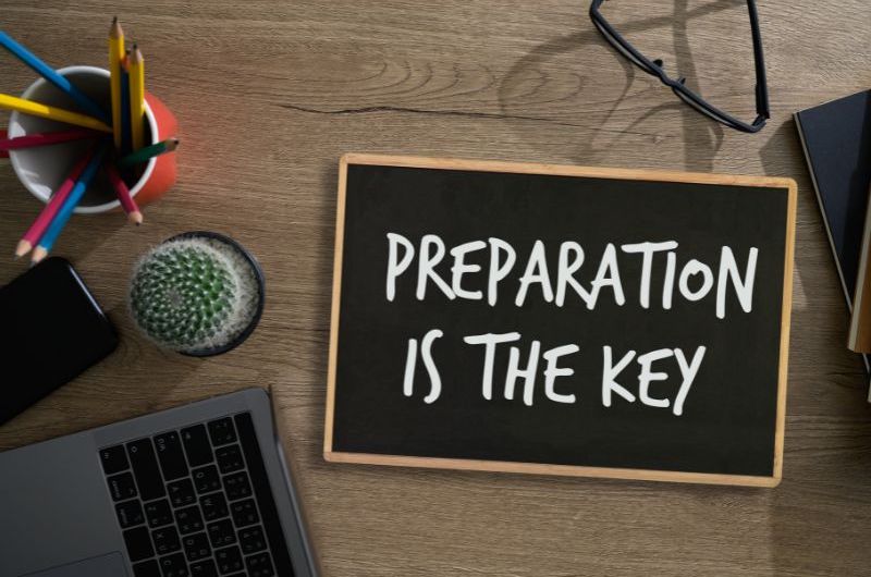 An image of a chalkboard with “PREPARATION IS THE KEY” written on it. The chalkboard is surrounded by glasses, a laptop, and a cup with colored pencils in it.