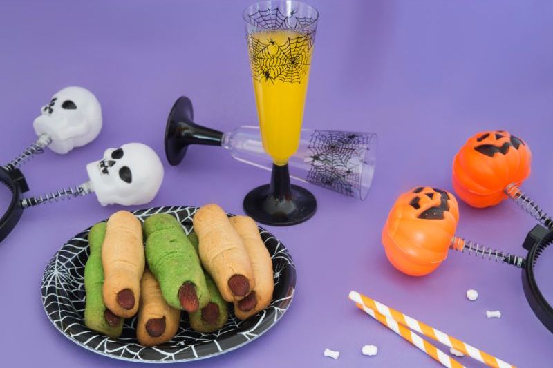 An image of various Halloween items. On a plate, there are corn dogs made to look like fingers with fingernails. There are also Halloween headbands and a glass of orange juice.