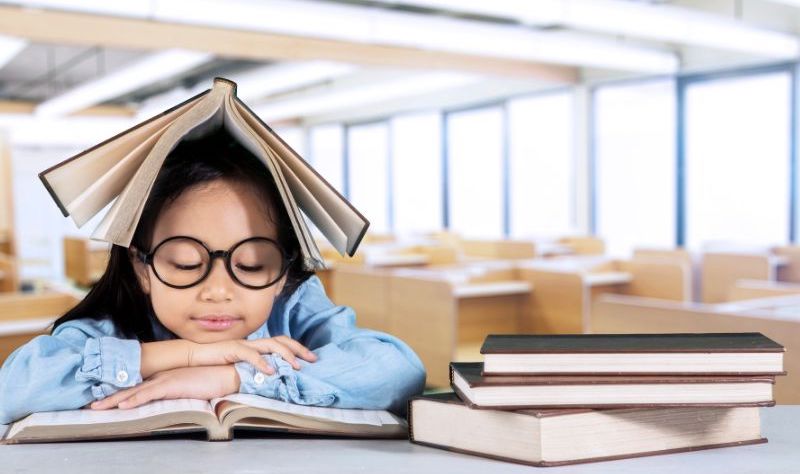 A young student has her head on the desk with a book open on top of her head and a stack of books beside her.